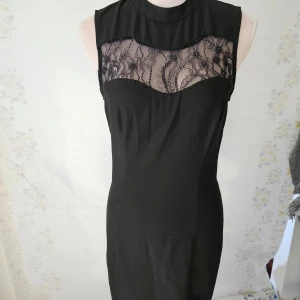 Svart ärmlös klänning med spetsdetaljer - En elegant svart ärmlös klänning med spetsdetaljer över bröstet och en hög hals. Klänningen har en figurnära passform och är tillverkad i ett mjukt material.