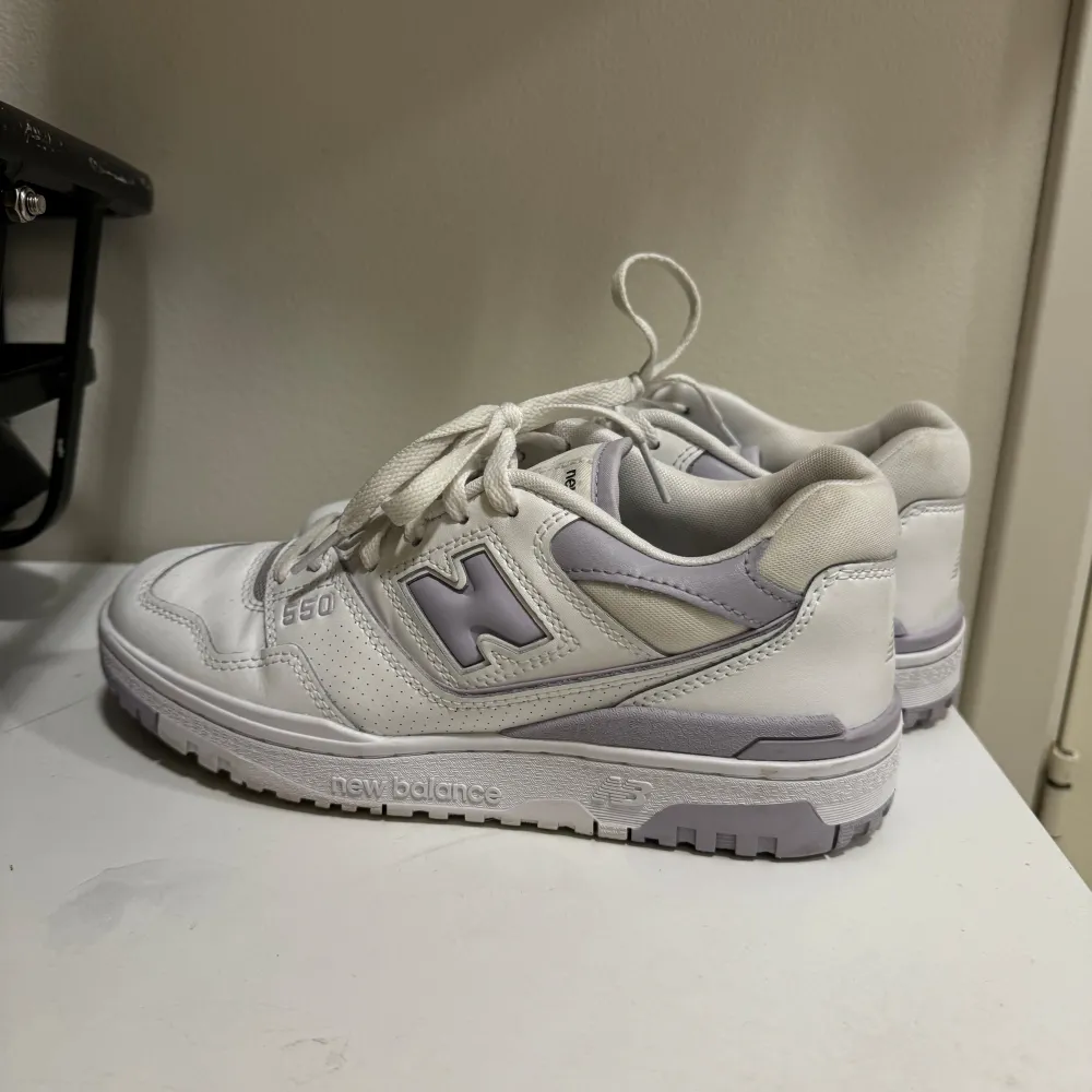 Ett par vita och ljuslila New Balance 550 sneakers med snörning. Skorna har en robust sula och är designade för komfort och stil.. Skor.