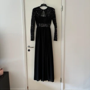 Svart långklänning med spetsdetaljer - En elegant svart långklänning med långa ärmar och spetsdetaljer. Klänningen har en öppen rygg och är tillverkad av ett lätt och flödande material som ger en vacker siluett. Storlek 34 men passar även 36. 