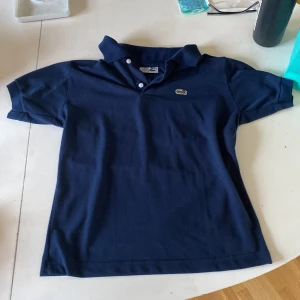 Lacoste Piké - Säljer en marin blå Lacoste piké som är utmärk till sommaren och våren som är i 9/10 skick. Ny kan kosta upp till 1100 kr.