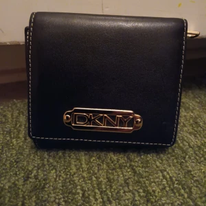 Svart och Guldig DKNY Plånbok - Denna DKNY plånboken är svart med detaljer i guld. Den har endast använts ett fåtal gånger och är i utmärkt skick.  Plånboken är 11 cm bred och 9,5 cm hög när den är stängd. När den är öppen är den ca 29,5 cm. 