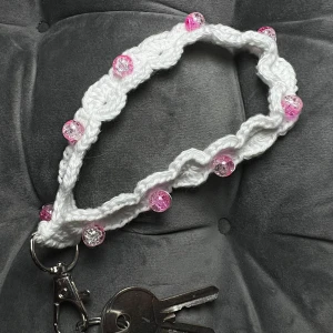 Nyckelband - Egenvirkat nyckelband i vitt bomullsgarn med fina rosa krackelerade pärlor och en karbinhake som avslut. 18 cm lång och ca 26 cm i omkrets. Passar perfekt på handleden eller i din ficka!