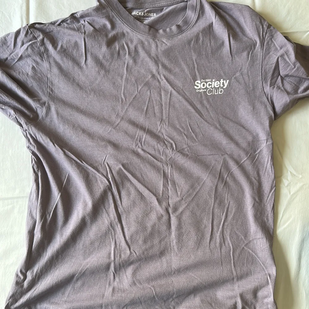 En lila t-shirt med Society originals club tryck på vänster bröst. T-shirts.