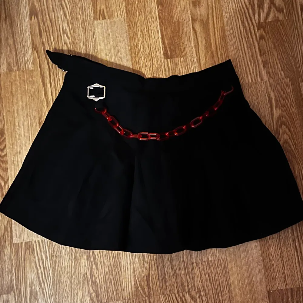 Svart kjol med en röd kedja i plast. Den är halv så ena sidan är öppen så tipsar om att ha ett par shorts under.. Kjolar.