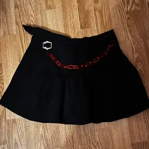 Svart kjol med en röd kedja i plast. Den är halv så ena sidan är öppen så tipsar om att ha ett par shorts under.