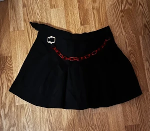 Svart kjol med en röd kedja - Svart kjol med en röd kedja i plast. Den är halv så ena sidan är öppen så tipsar om att ha ett par shorts under.