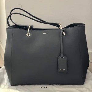 DKNY VÄSKA - DKNY - väska  Färg grå utsida med svart insida.  3 innefack.  Magnet lås.  Använd 1-2 gånger så är i skick som helt oanvänd.   