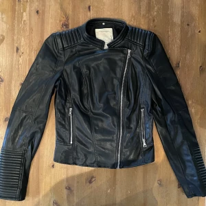 Skinnjacka - Lite sliten på vissa ställen men det syns knappt när man har på sig den. Två fickor med dragkedja samt dragkedja vid ärmarnas öppningar. Inte äkta skinn.