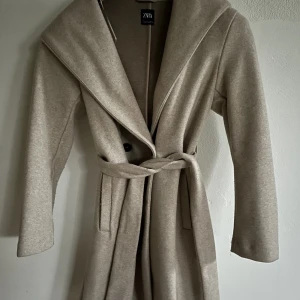 Superfin kappa - Säljer denna superfina kappa från zara, perfekt nu till hösten. Medellång beige kappa, men luva bak och avtagbart skärp/rep. Använd ett fåtal gånger. Säljer för 400kr + köparen står för frakten