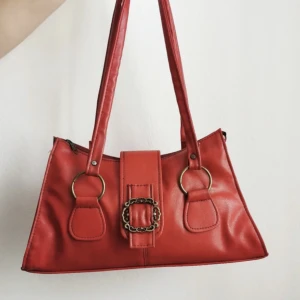 Röd handväska med gulddetaljer - Säljer denna klarröda, rymliga vintageväska i läderimitation.  Bredd nedtill: 37 Bredd upptill: 26 Djup: 9 Längd på band: ca 64 Höjd: 19  Bra skick men lätt missfärgad runt fästena till banden, se närbild. Borde gå att tvätta!  Bilder från förra säljaren