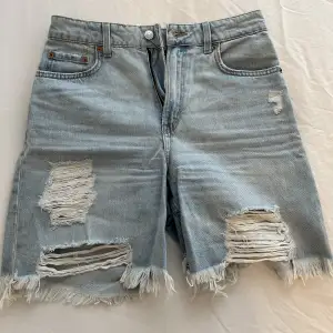 Ljusblåa slitna jeansshort. De slutar precis ovanför knäna och är oanvända. 