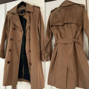 GANT Trenchcoat - I fint skick med läderdetaljer och grönrutigt innerfoder. Slutar vid knäna på mig som är 172 cm lång. Nypris ca 3500 kr.