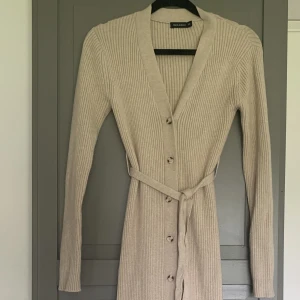 Ribbstickad klänning  - Lång ribb stickad klänning i en beige melange färg. Klänningen har långa ärmar knappar hela vägen fram och ett stickat bälte i midjan.  Klänningen sträcker sig ner till vaden.