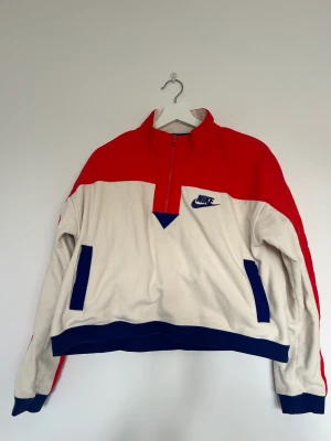 Nike fleecetröja  - En croppad fleecetröja från Nike. Lila detaljer i bomull och orange i ”vindjackematerial”. Storlek L passar unisex. Tröjan har en ficka på framsidan med två ingångar. Använd fåtal gånger. Mycket fint skick!