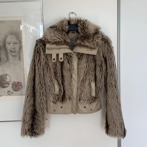 Kort pälsjacka - Kort ljusbrun pälsjacka med läderdetaljer. Fuskpäls, pälsen är mjuk. Vadderad inifrån. Storleken är L men skulle säga att den snarare är en M, stor S, beror på hur tjock tröja man har under. Väldigt fin jacka som sitter snyggt! Skriv för frågor/fler bilde