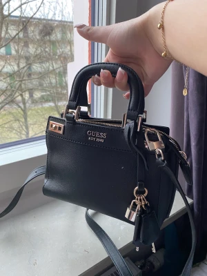 Svart Guess väska  - Snygg svart guess väska med guld detalj. Använd några gånger. Tillkommer även ett längre band till väskan.