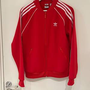 Röd Adidas sweat jacket med vita ränder i storlek 38. Har två fickor på framsidan med dragkedja. Är använd fåtal gånger, är som ny. Nypris 600 kr. 