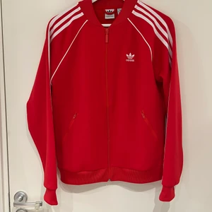 Röd Adidas sweat jacket strl 38  - Röd Adidas sweat jacket med vita ränder i storlek 38. Har två fickor på framsidan med dragkedja. Är använd fåtal gånger, är som ny. Nypris 600 kr. 