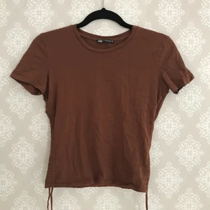 Brun topp / t-shirt - Brun topp från Zara (ca 2020) med justerbar "ruchning" samt knytning. Användes flitigt ett tag men inga tydliga defekter och i bra skick🤎 Hör av dig om du vill ha fler bilder eller några frågor t.ex. mått eller annat. Plagget är ej stryket i denna bilden.
