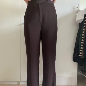 36 - Mörkbruna kostymbyxor i lite glansigt material. Köpta ifrån Beyond Retro så osäker på både material och storlek. Passar mig som är en 36/38 i midjan samt 170cm lång (går ner till fötterna). Raka i modellen. Nyskick! Köpare står för ev frakt🤎