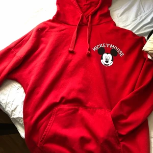 Röd hoodie i storlek M. - Röd hoodie med ett litet Musse Pigg-tryck på framsidan och ett stort på ryggen. Har resår i midjan och ärmarna, luva med snören och en ficka. Är i storlek M, men passar även L.