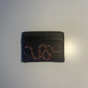 Gucci Plånbok (Kingsnake print GG Supreme card case) - Orignalpris på plånboken är 2468 kr och därför ligger denna på 900 kr. Plånboken har använts i ett halvår och är i gott skick, finns några få skråmor på vissa ställen. Men de syns knappt. Plånboken är 10 cm bred och 7 cm hög. Den är gjord i svart läder.