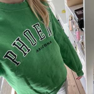 En grön sweatshirt med texten ”phoenix”.  Den är från ginatricot och är i storlek XS. 