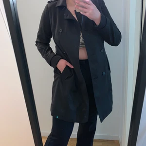Svart trenchcoat från VILA strl M - Här har vi en riktigt fin trenchcoat från VILA i strl M. !HELT OANVÄND! enbart testad. Perfekt nu till hösten ju. Jag är 169cm och den slutar ungefär på mitten av mina lår. Nypris är 599kr 
