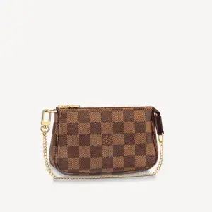 The Mini Pochette Accessoires in Damier Ebene canvas.  Denna väska är nästan alltid slutsåld i butik och nätet, väldigt svår att få tag på. Kartong, dustbag och kvitto finns.  Its supple, rounded shape gives this pouch a particularly pleasant feel, while its zipped compartment offers a surprising amount of space. It can be hand-carried or attached to a larger bag thanks to the gold-tone chain and hook.   Priset kan diskuteras lite