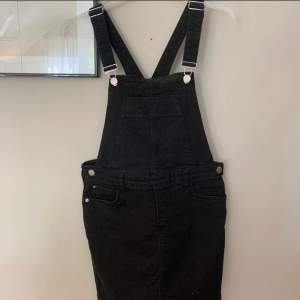 En svart hängselklänning i jeans material från River island i storlek M, men den är ganska liten för sin storlek! Klänningen har en bröstficka och två bakfickor, en kortare modell!  Frakt tillkommer på 79kr!