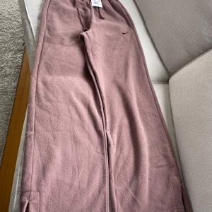 Nike mjukisbyxor i smokey mauve - Mysiga och breda mjukisbyxor från Nike i färgen smokey mauve. Byxorna har resår i midjan och en liten Nike-logga på ena benet. Perfekta för en avslappnad stil och sköna att bära hela dagen. Nyskick med prisetikett kvar