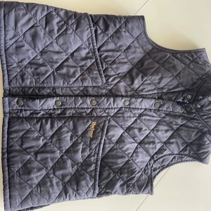 Mörkblå quiltad väst från Barbour - Säljer en  mörkblå quiltad väst från Barbour med tryckknappar framtill och två stora fickor. Klassisk och stilren design med Barbour-logga broderad på fickan. Perfekt för lager-på-lager och passar till många olika outfits.