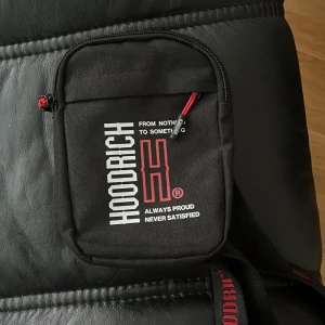 Svart axelväska från Hoodrich - Säljer en svart axelväska från Hoodrich med vit logga och text på framsidan. Väskan har en dragkedja upptill och en extra ficka framtill med röd detalj på dragkedjan. Perfekt för att bära med sig det viktigaste.