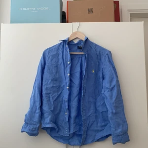 Blå linneskjorta från Polo Ralph Lauren - Snygg blå skjorta i linne från Polo Ralph Lauren med klassisk gul broderad logga på bröstet. Skjortan har lång ärm, knappar framtill och en avslappnad passform. Perfekt för en stilren och fräsch look. Storlek L barn, storlek XS vuxen. Kom dm vid funderingar!