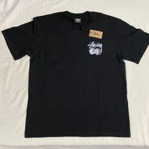 Svart t-shirt med tärningsmotiv från Stüssy - Svart t-shirt från Stüssy med stort vitt logotryck och tärningar på både bröstet och ryggen. Klassisk passform och rund halsringning. Perfekt för dig som gillar streetwear och vill sticka ut med en ikonisk design.