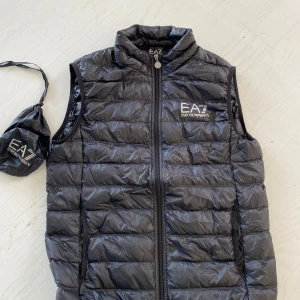 Svart glansig väst Emporio Armani EA7 - Snygg svart väst från Emporio Armani EA7 skick 9,9/10med dragkedja framtill. Västen är i väldigt bra skick  och levereras med en praktisk förvaringspåse. Västen är använd max 3 gånger och ser ut som att den är ny från butik. Säljer pga har vuxit ur den. Skriv för frågor
