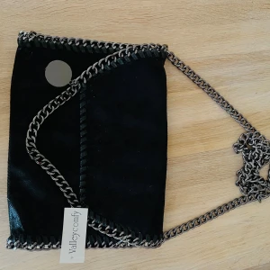 Svart axelväska med kedjedetaljer från Valleycomfy - Snygg svart axelväska med silverfärgade kedjor längs kanterna och som axelrem. Väskan har ett leopardmönstrat innerfoder och en innerficka med dragkedja. Perfekt för att lyfta din outfit.