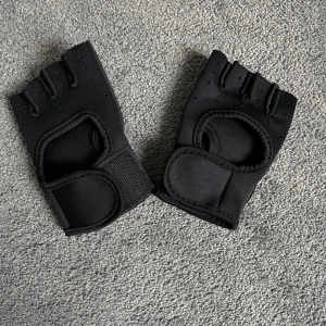 Svarta träningshandskar med korta fingrar - Svarta träningshandskar med korta fingrar och justerbar kardborre vid handleden. Handskarna har ventilerande material och förstärkta partier i handflatan för extra grepp. Perfekta för gym eller styrketräning.