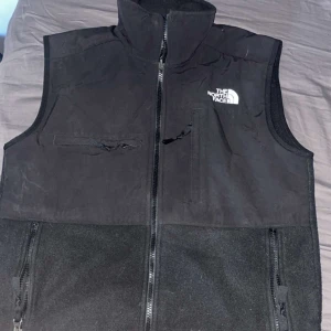 Svart fleeceväst från The North Face - Säljer en svart fleeceväst från The North Face med dragkedja framtill och flera fickor. Västen har hög krage och logga på både fram- och baksidan. Perfekt för lager-på-lager under kyliga dagar.