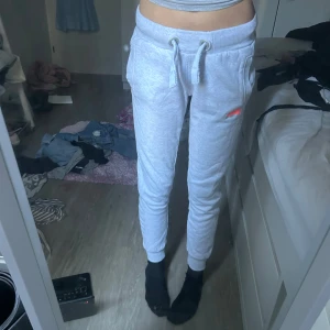 Grå mjukisbyxor från Superdry - Gråa mjukisbyxor från Superdry med dragsko i midjan och broderad logga i orange på ena benet. Slim fit-modell med muddar vid bensluten och mjukt bomullsmaterial. Perfekta för en avslappnad stil.