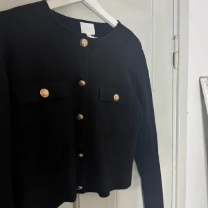 Svart cardigan jacka med guldknappar - Säljer en svart cardigan jacka med snygga guldknappar framtill och två fickor på bröstet. Jackan har rund halsringning och lång ärm, perfekt för att lyfta din outfit med en klassisk touch.