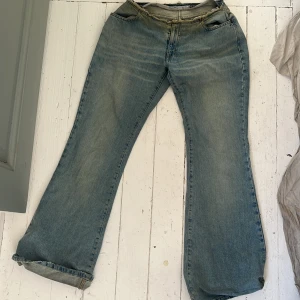 Blå bootcut jeans med patch - Säljer ett par blå bootcut jeans med en mörkblå patch på baksidan och slitningar vid ena bakfickan. Jeansen har klassisk femficksdesign och låg midja. Perfekta för dig som gillar en avslappnad och retro stil. Glitter runt midjan. 
