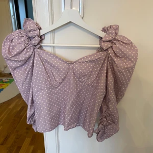 Ljuslila prickig blus med puffärmar och rosetter - Superfin ljuslila blus med vita prickar och stora puffärmar med rosetter på axlarna. Blusen har markerad byst, dragkedja i ryggen och är tillverkad i ett mjukt bomullsliknande material. Perfekt för dig som vill sticka ut med en söt och trendig topp!