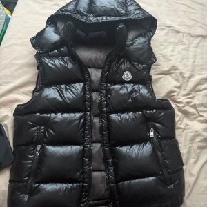 Svart väst från Moncler med huva - Säljer en svart, glansig dunväst från Moncler med huva och dragkedja. Såklart äkta Västen har två fickor med dragkedja framtill och Moncler-logga på bröstet. Perfekt för lager-på-lager och snygg till många outfits. Stolek 2 och sitter som S pris går att diskutera 