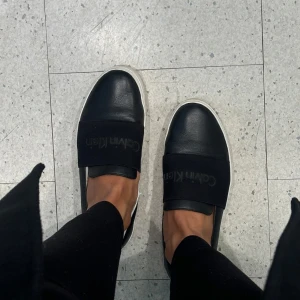 Svarta slip-on sneakers från Calvin Klein - Säljer ett par svarta slip-on sneakers från Calvin Klein med bred resår över foten där loggan syns. Skorna har vit sula och är i ett stilrent, minimalistiskt utförande. Perfekta för en clean och modern look. Boxen finns med