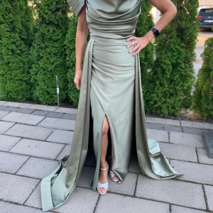 Ljusgrön långklänning i satin med slits - Säljer en elegant ljusgrön långklänning i satin med draperad överdel och hög slits framtill. Klänningen har en axel och lång släp baktill, vilket ger en lyxig känsla. Perfekt för bal eller fest. Snygg passform och vackert fallande material.