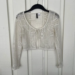 Vit spetsig croppad bolerotopp från H&M Divided - Säljer en vit, croppad bolerotopp från H&M Divided. Toppen är i spets med lång ärm och knytning framtill. Perfekt att styla över ett linne eller klänning för en trendig look. Den är använd några gånger men är ju jättebra skick. Det är ljudet som gör att den ser lite missfärgad ut, men den är vit i verkligheten! Skriv om du har några frågor!💕