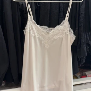 Vit spetslinne med smala axelband - Säljer ett vitt linne med tunna axelband och vacker spetsdetalj längs urringningen. Perfekt att styla under en kavaj eller till jeans för en enkel men snygg look.