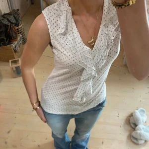 Vit prickig blus med volangdetaljer - Säljer en söt vit blus med små svarta prickar och volangdetaljer framtill. Blusen är ärmlös och har en v-ringad design som ger en elegant touch. Perfekt för en stilren look!