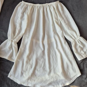 Vit offshoulder tunkia med bälte från Boohoo - Säljer en vit offshoulder tunika från Boohoo med långa ärmar och volangdetaljer vid ärmsluten. Klänningen har ett matchande bälte i midjan och är tillverkad i ett lätt, luftigt material. Perfekt för varma dagar eller sommarens tillfällen.